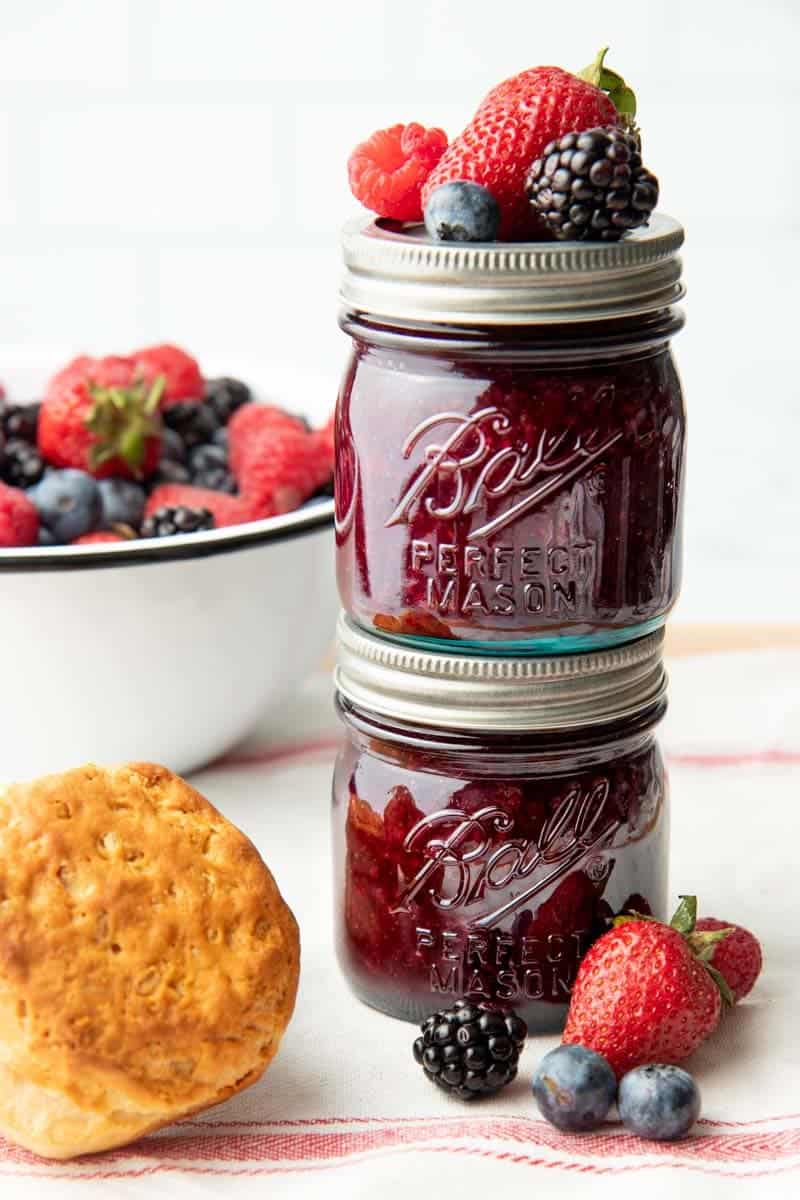 Two vintage Ball jars filled with mixed berry jam stacked with fresh berries on top, beside a bowl of mixed berries and a fluffy biscuit.