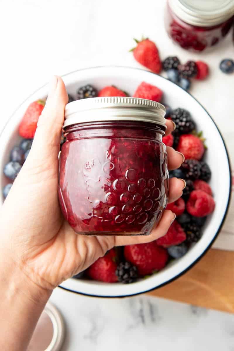 Close-up of a hand holding a finished jar of jam over a bowl of fresh berries.