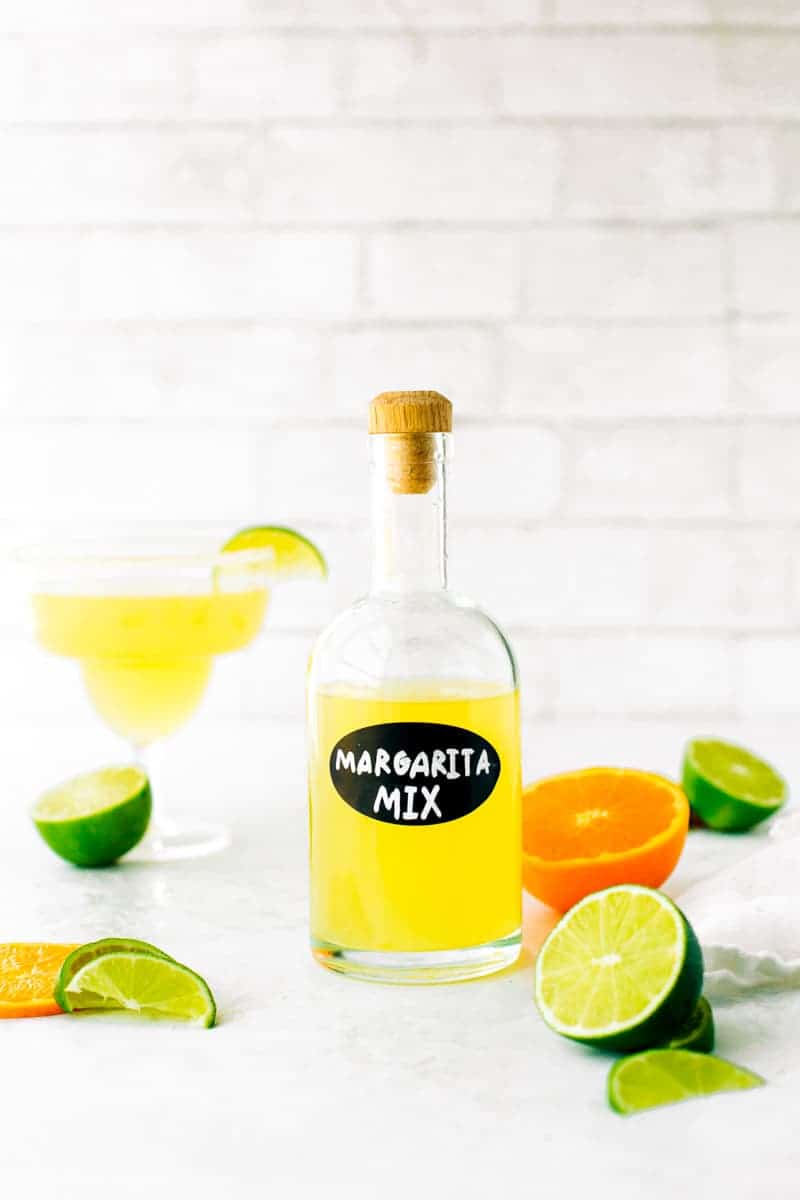 Homemade margarita mix in a glass bottle surrounded by fresh cut limes and oranges.