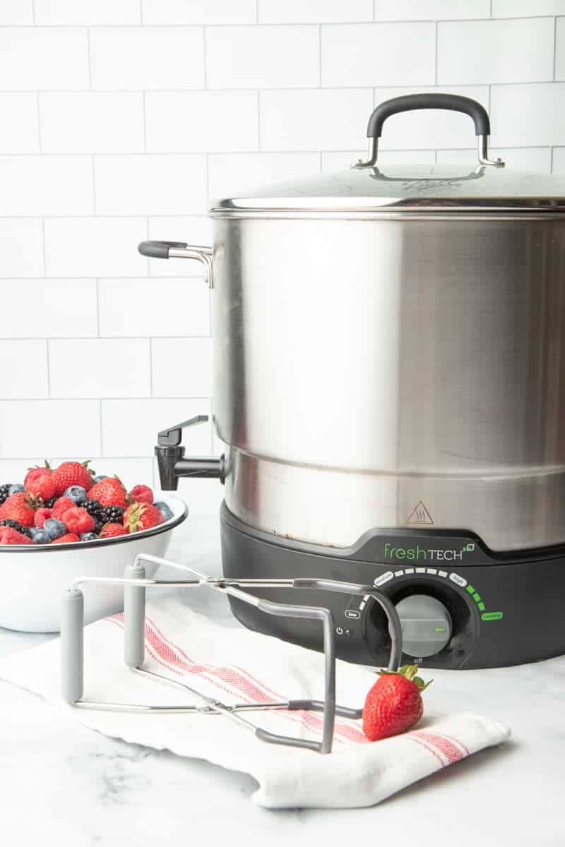 FreshTech water bath canner sitting on a kitchen counter with jar lifters and fresh berries beside it.