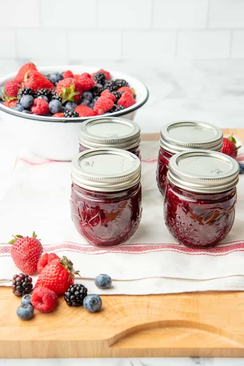Four Ball jars of finished mixed berry agave jam cooling on a towel with a bowl of fresh mixed berries in the background.