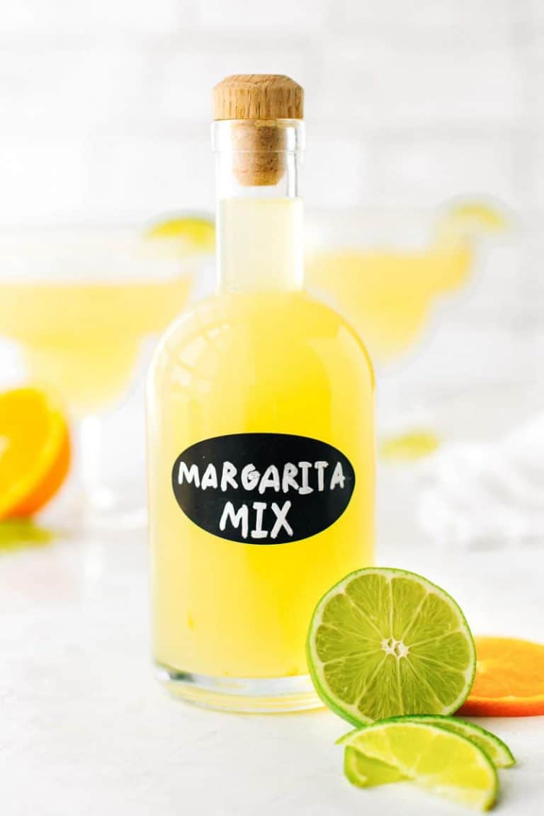 Close-up of homemade margarita mix in a glass bottle with a handwritten label and fresh cut limes and oranges.