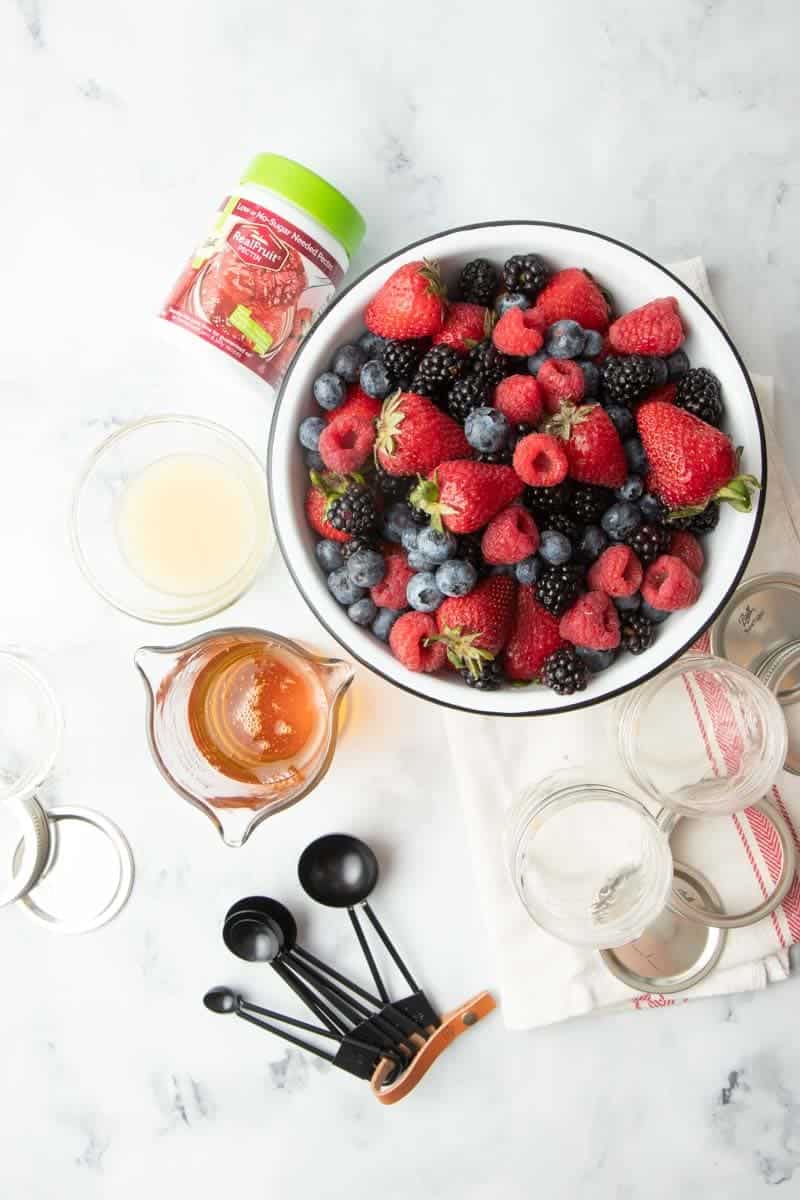 Overhead of ingredients needed for mixed berry agave jam recipe alongside tools such as measuring spoons and canning jars and lids.