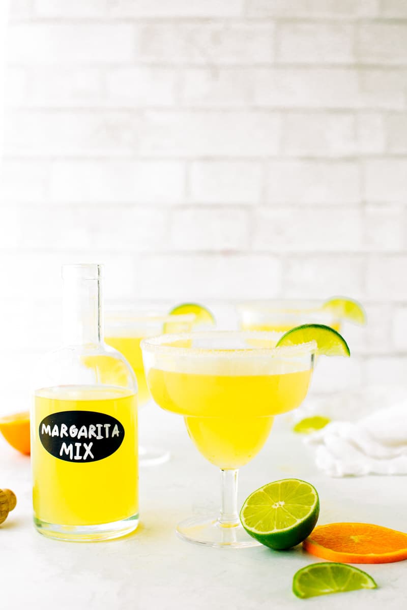 Bottle of homemade margarita mix next to margarita with a salt rim and fresh citrus.