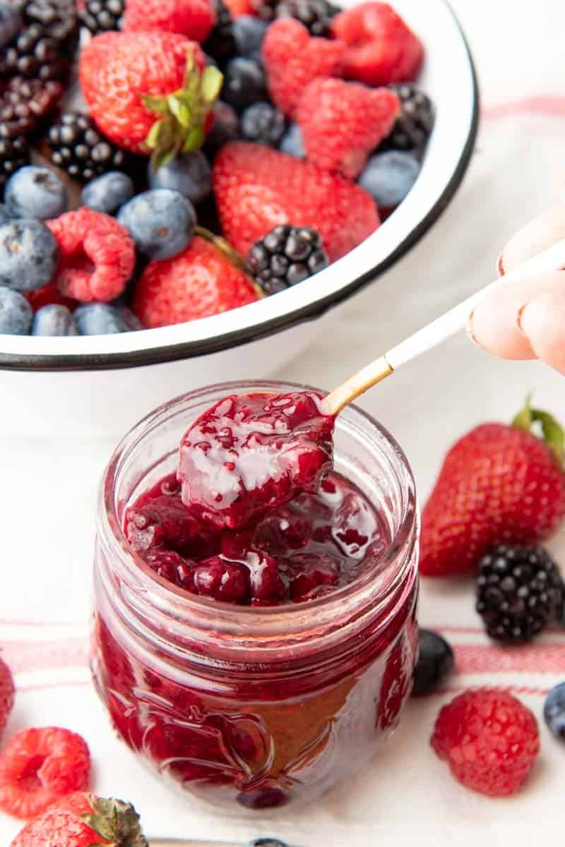 Scooping a spoonful of jam out of a full canning jar, with fresh mixed berries in a bowl in the background.