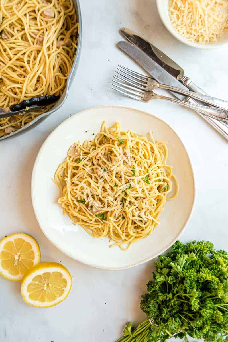 Overhead of spaghetti with canned clams in a bowl beside silverware and fresh garnishes.