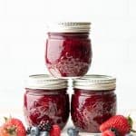 Three jars filled with mixed berry agave jam stacked in a pyramid with fresh berries in front.