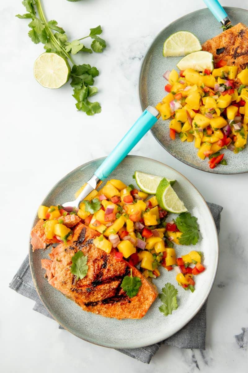 Overhead of two plates of grilled salmon with mango salsa beside fresh lime and cilantro.