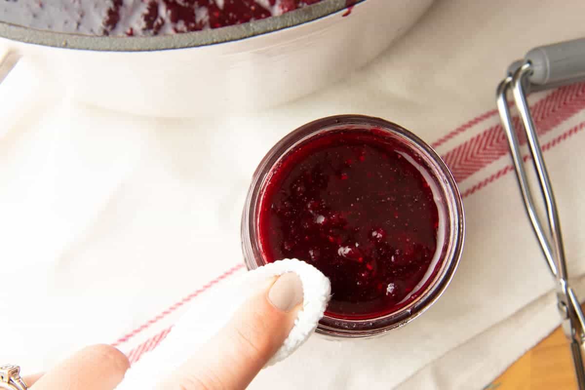 Close-up of clean cloth wiping rim of canning jar to remove excess jam after filling.