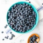 Overhead of fresh blueberries in a large bowl with a wooden spoon filled with blueberries alongside.