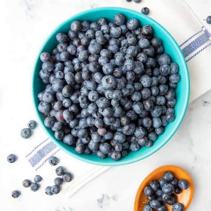 Overhead of fresh blueberries in a large bowl with a wooden spoon filled with blueberries alongside.