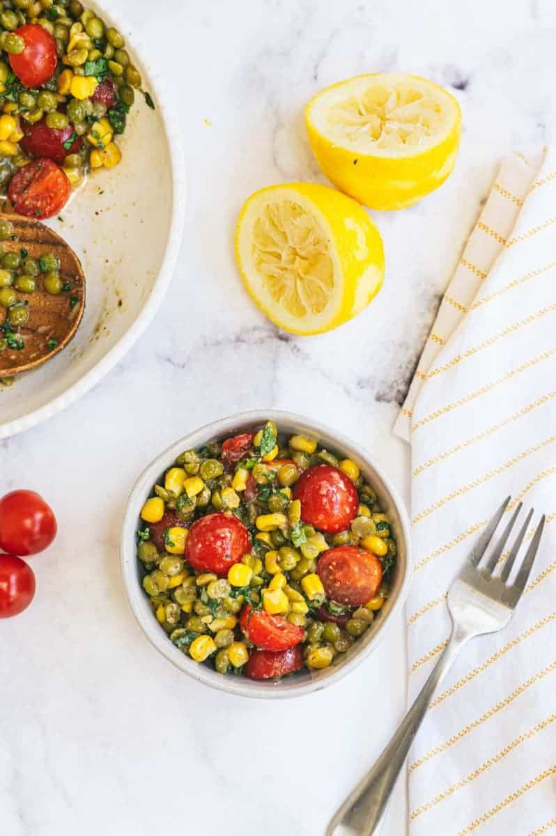 A single serving of summer split pea salad in a bowl with a fork alongside the larger serving bowl, fresh tomatoes, and zested and juiced lemon halves.