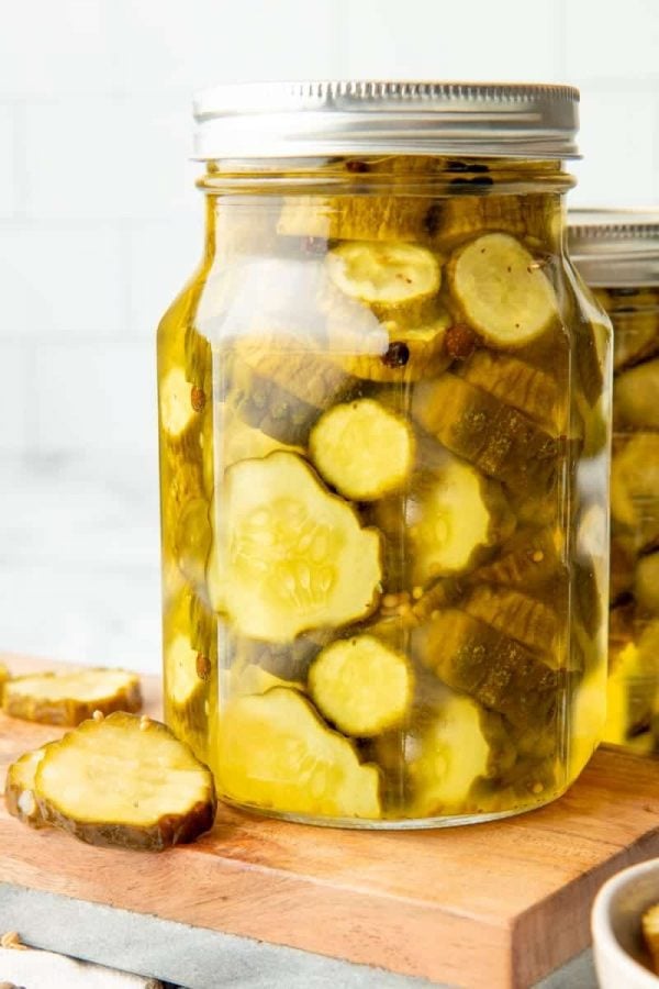 Close-up of full quart jar of canned bread and butter pickles on a wooden cutting board with fresh pickles beside it.