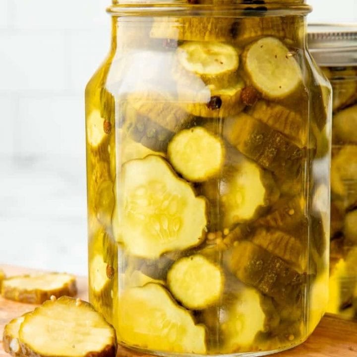 Close-up of full quart jar of canned bread and butter pickles on a wooden cutting board with fresh pickles beside it.