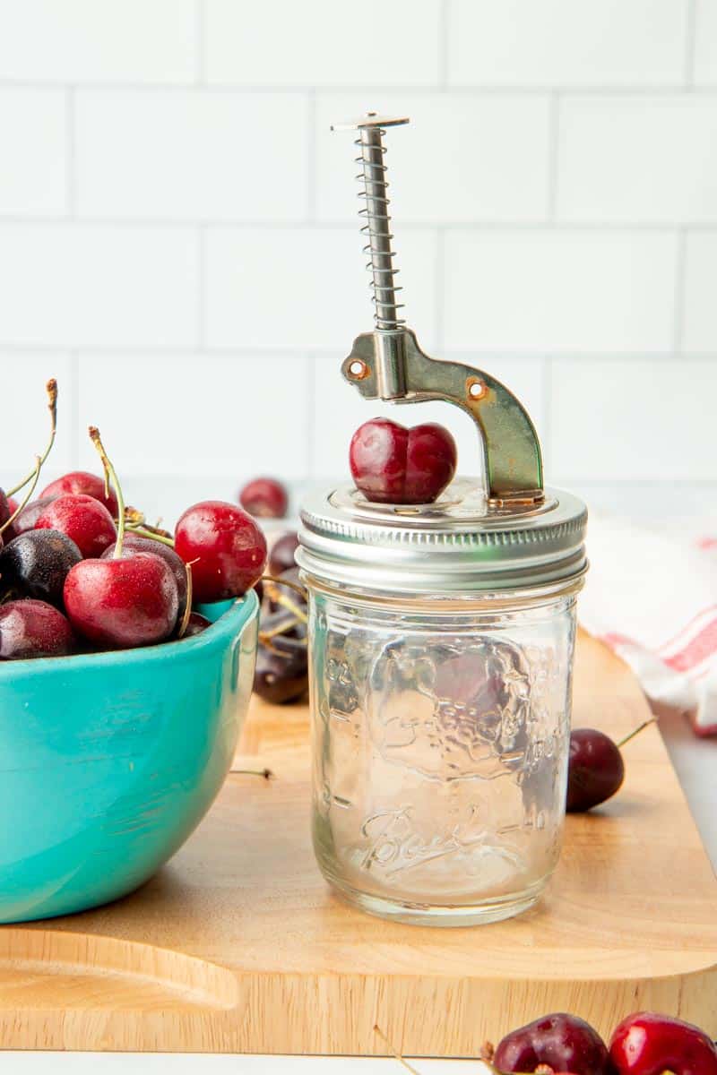 Mason jar push button cherry pitter with a cherry on it ready to be pitted, next to a bowl of fresh, sweet cherries.