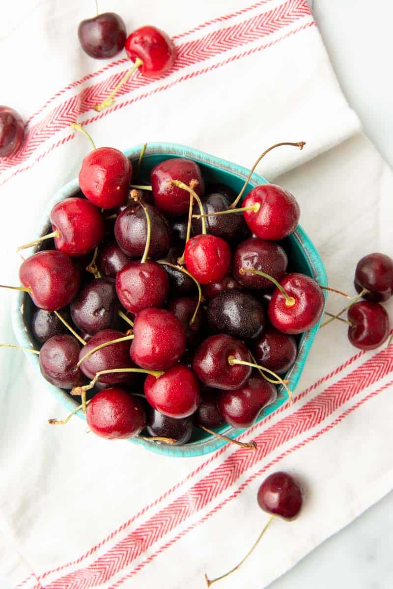 Overhead of fresh cherries spilled onto a clean kitchen linen from an overflowing bowl.