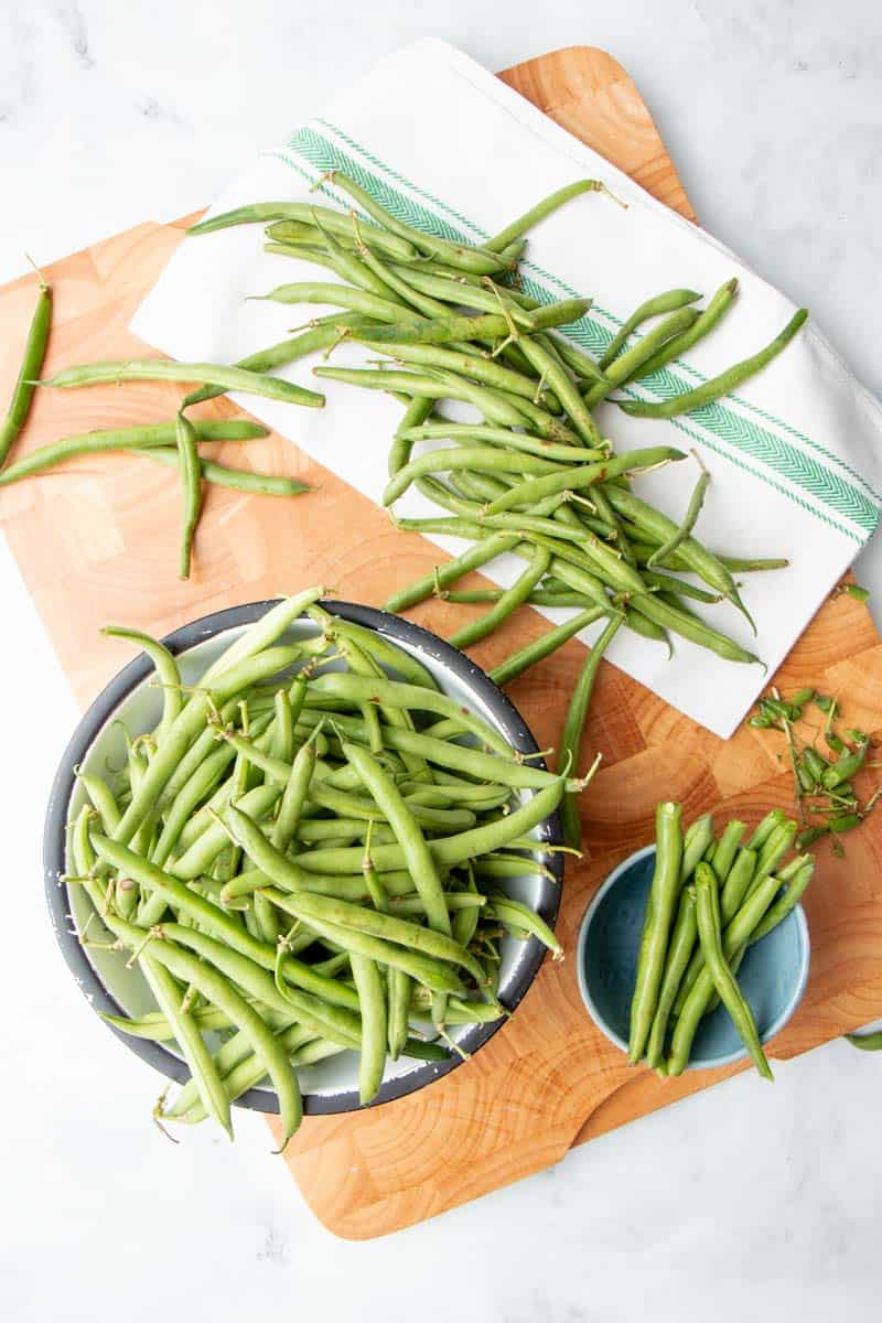 Overhead of fresh green beans in a bowl on a wooden cutting board alongside freshly snapped beans in another bowl.