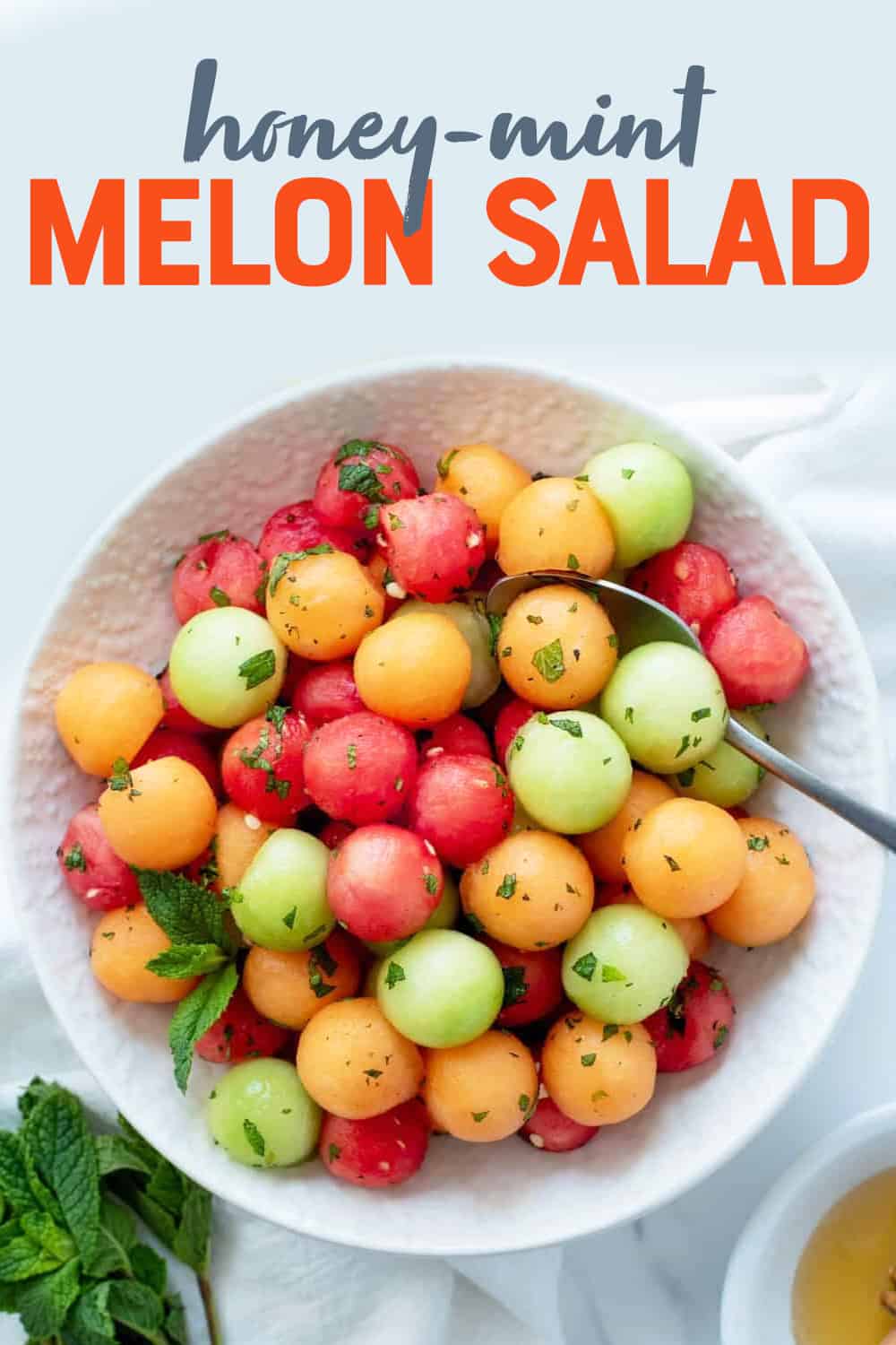 Overhead of finished melon ball salad in a serving bowl with fresh mint and honey. A text overlay reads, "Honey-Mint Melon Salad."