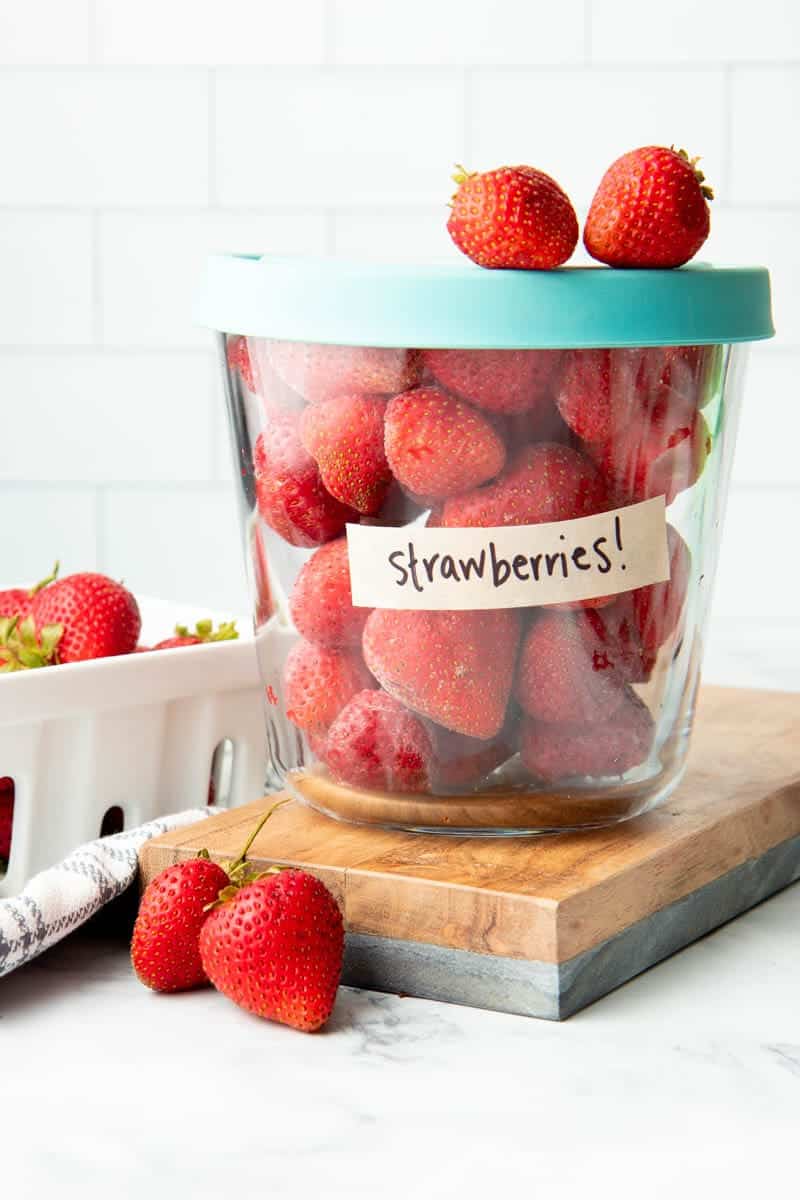 Lidded glass container labeled strawberries filled with whole, frozen strawberries sits atop a wooden cutting board alongside fresh strawberries.