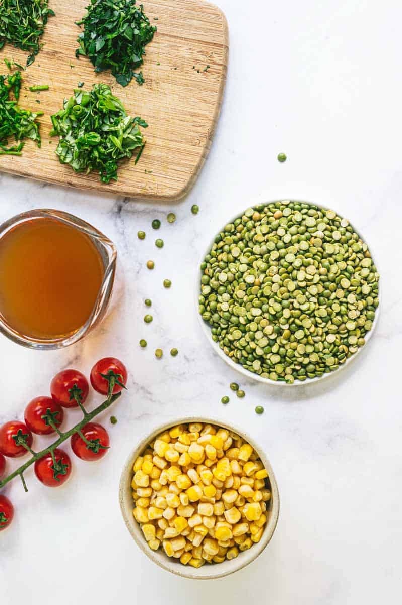 Ingredients for salad such as uncooked split peas, broth, frozen corn kernels, ripe cherry tomatoes on the vine, and chopped fresh herbs.