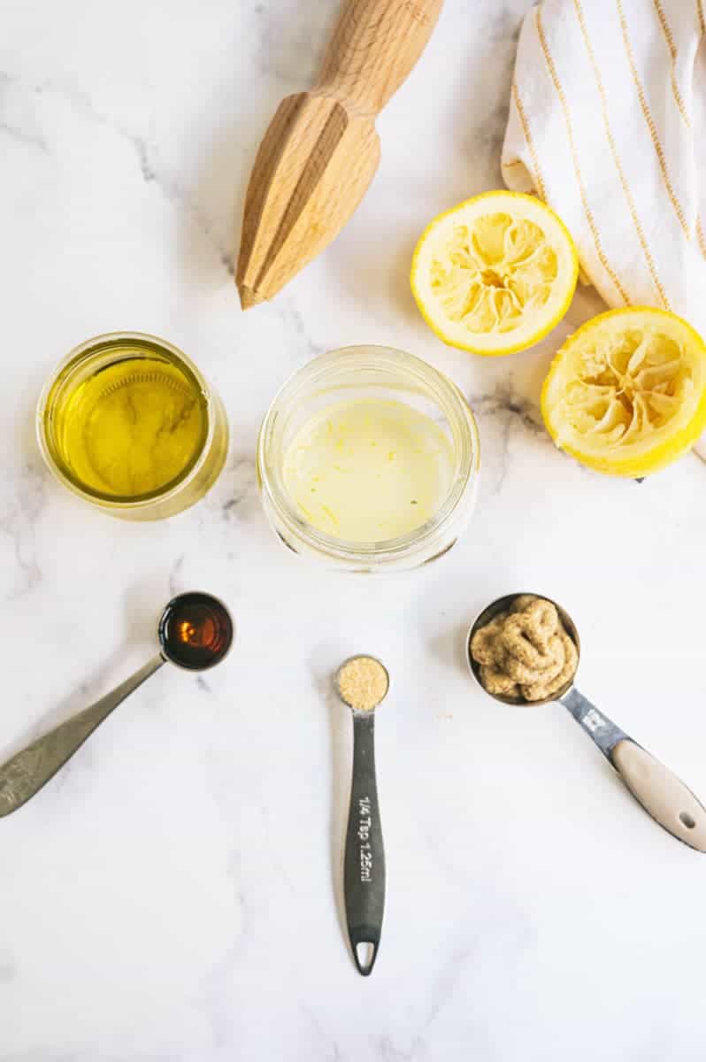 Ingredients for salad dressing set out in measuring spoons and cups alongside juiced lemon halves and wooden citrus reamer.