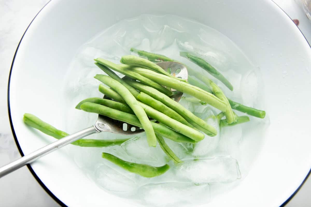 Overhead of a slotted spoon lowering blanched green beans into an ice bath.