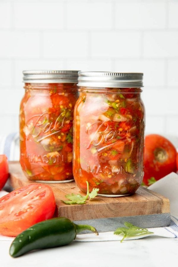 Two pint-sized Ball jars filled with canning salsa sit on a wooden cutting board with fresh cilantro, jalapeño, and tomatoes around them.