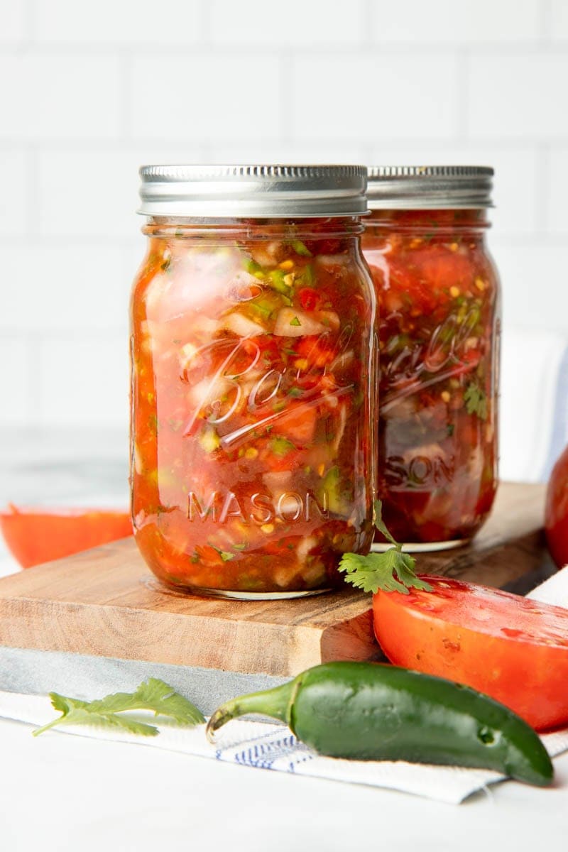 Two pint jars of zesty salsa sit on a cutting board with fresh jalapeño, tomato, and cilantro beside it.