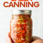Close-up of a jar of salsa cupped into the palm of someone's hand. A text overlay reads, "Step-by-Step Salsa Canning."