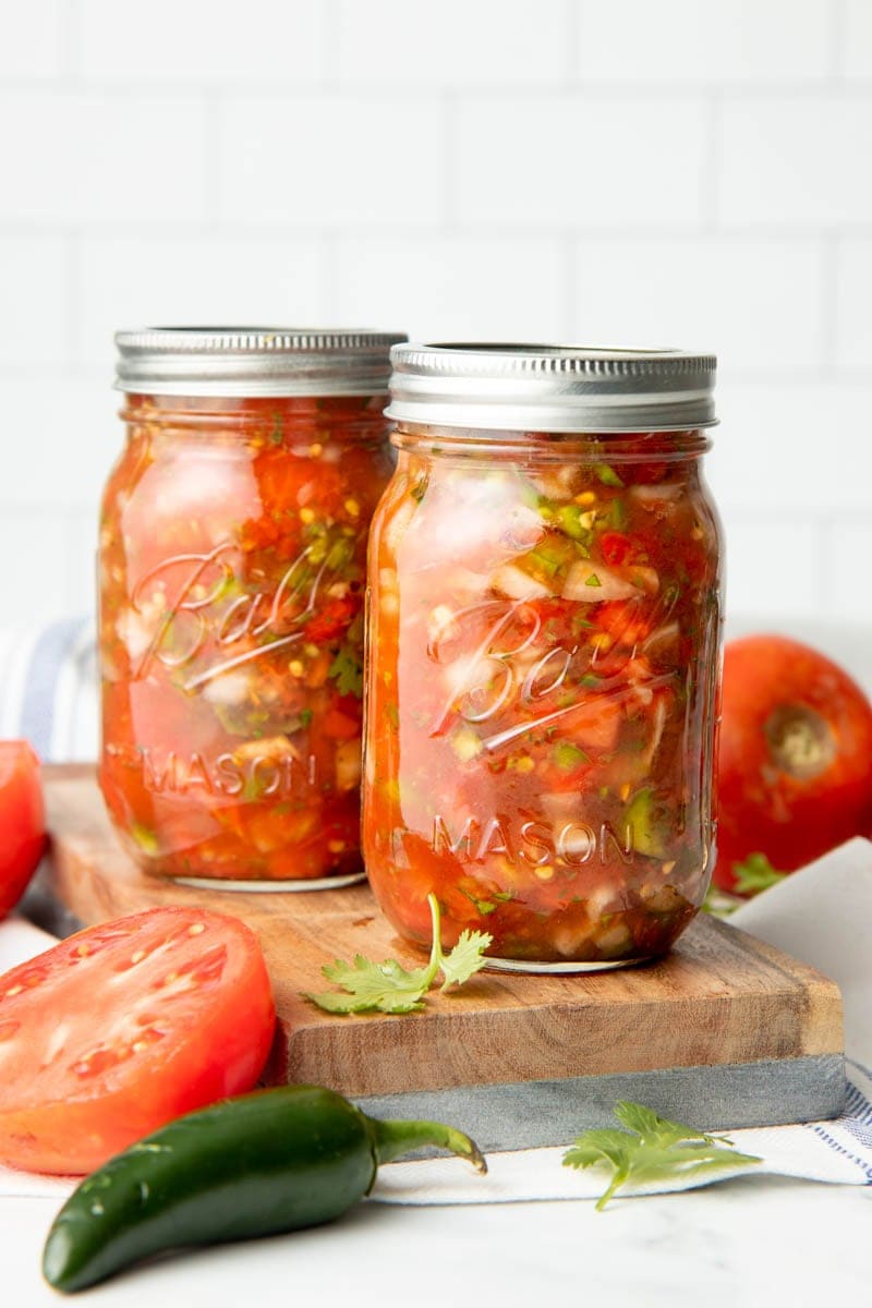 Two mason jars filled with canning salsa on a wooden cutting board surrounded by fresh ingredients.