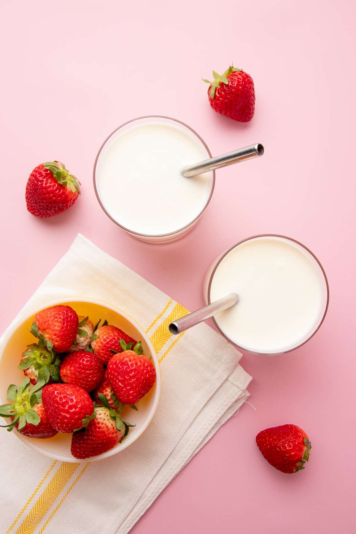 Overhead of two full rocks glasses of homemade kefir with metal straws alongside a bowl of whole, fresh strawberries.