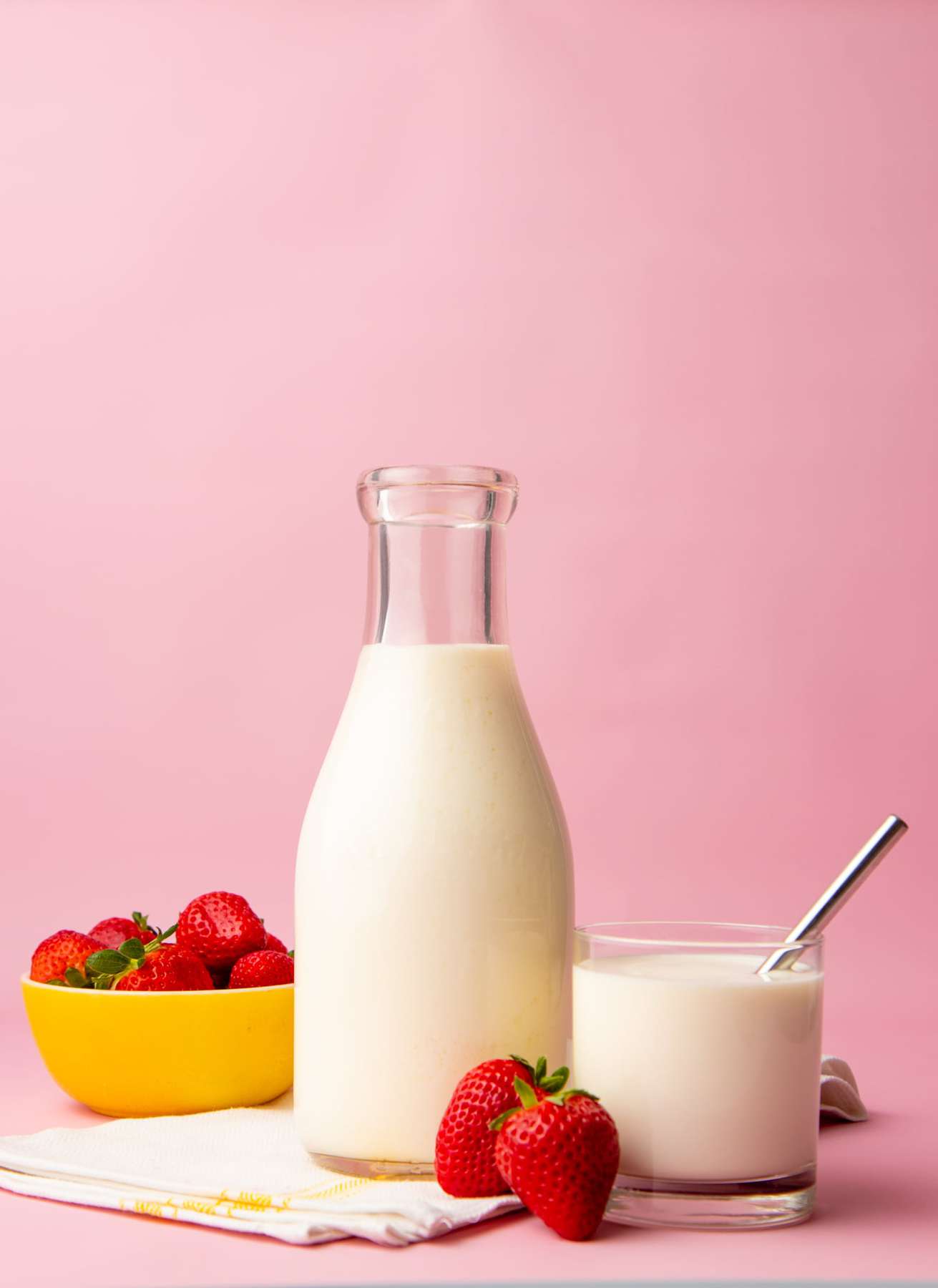 A glass carafe filled with homemade kefir sits on a linen kitchen towel with a full tumbler and a bowl of fresh strawberries on either side of it.