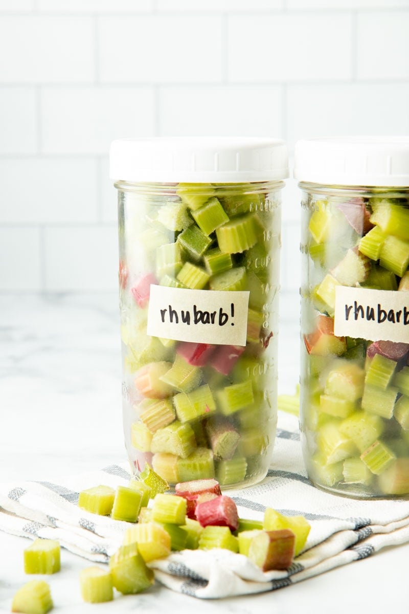 Close-up of two mason jars labeled and filled with individually frozen pieces of cut rhubarb, standing up on a kitchen towel.