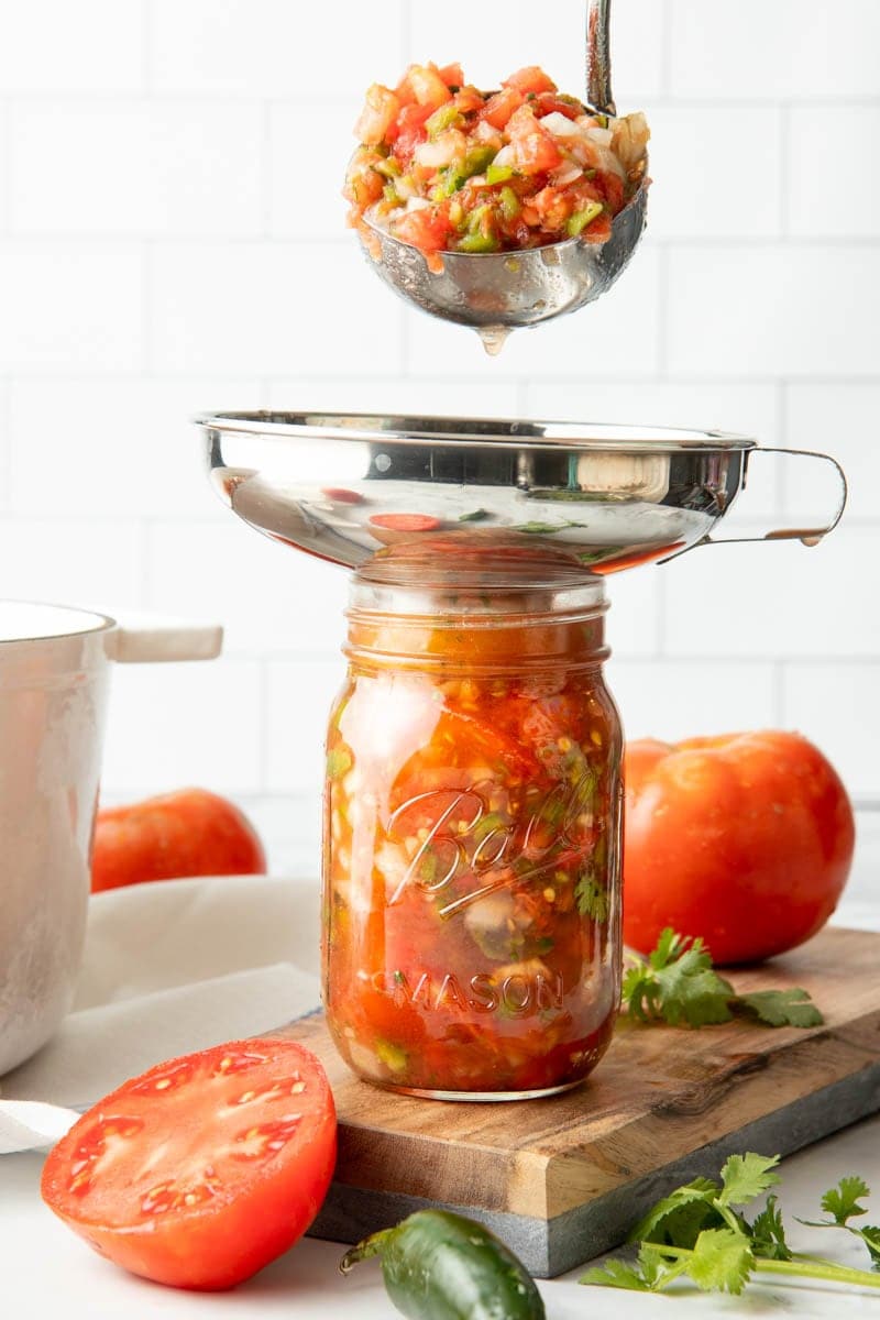 Canning salsa in progress: A ladle full of salsa hovers over a mason jar with a canning funnel in it.