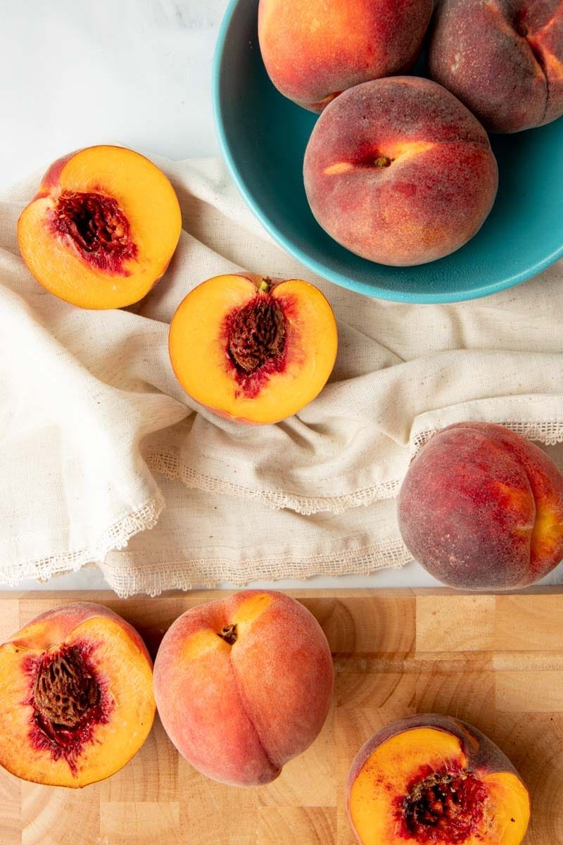 Overhead of whole and halved fresh peaches on kitchen linen and a wooden cutting board.