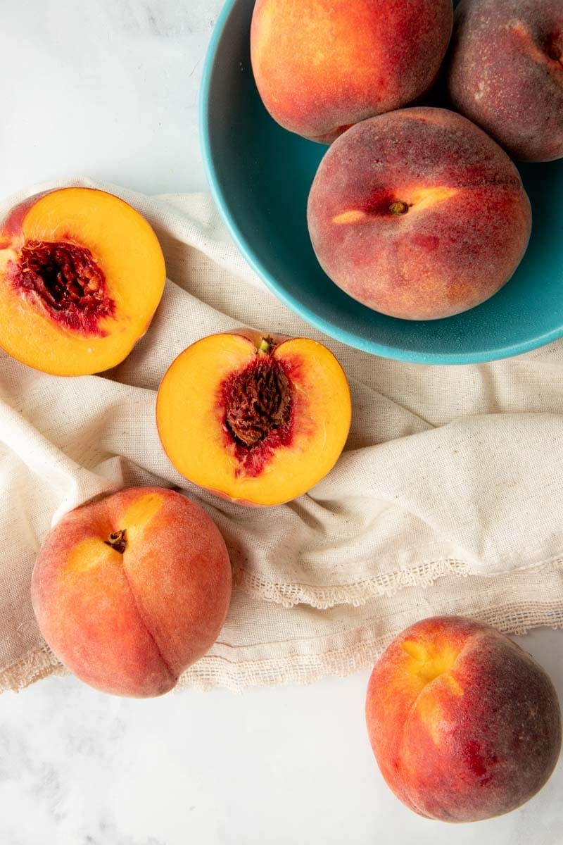 Overhead of whole, fresh peaches in a bowl and halved peaches on kitchen linen.
