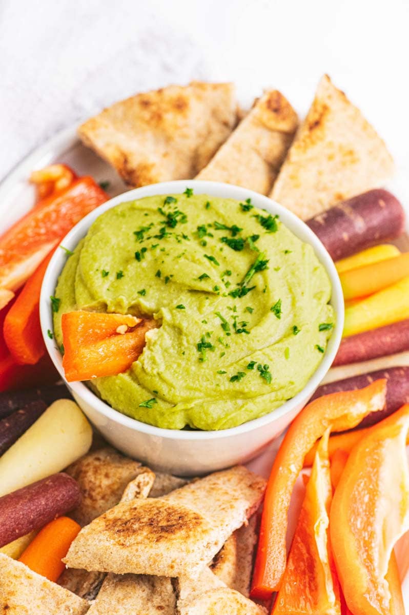 Close-up of a red bell pepper strip dipped into a bowl of split pea hummus on a party platter.
