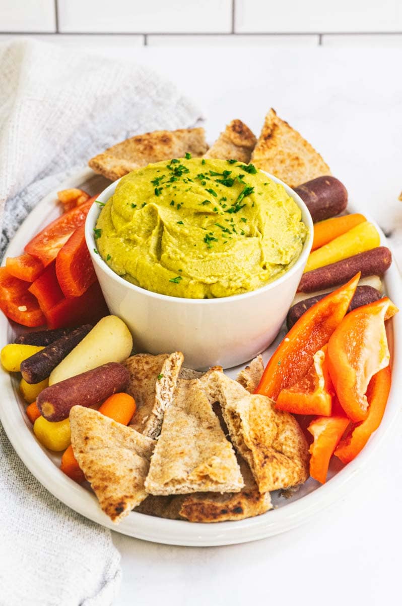 A large bowl of split pea hummus sits in the center of a platter of pita chips and fresh vegetables for dipping.