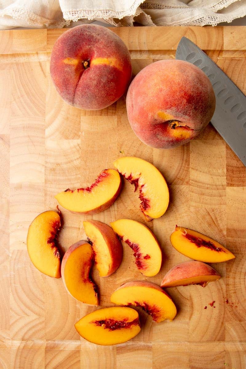 Overhead of fresh peach slices on a wooden cutting board with two whole peaches and a knife.