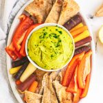 Overhead of a party platter with a bowl of split pea hummus in the center surrounded by pita chips, rainbow carrot sticks, and red bell pepper strips.