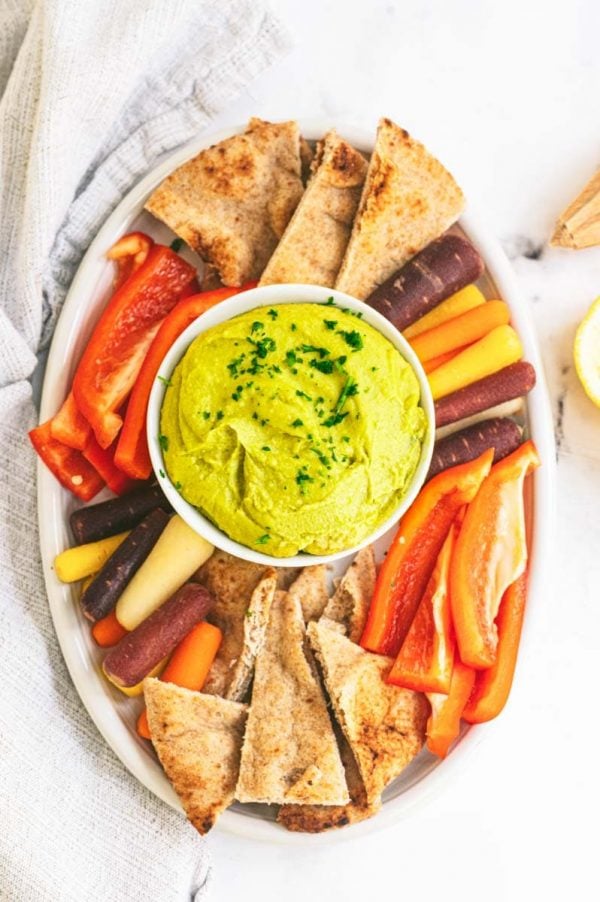 Overhead of a party platter with a bowl of split pea hummus in the center surrounded by pita chips, rainbow carrot sticks, and red bell pepper strips.