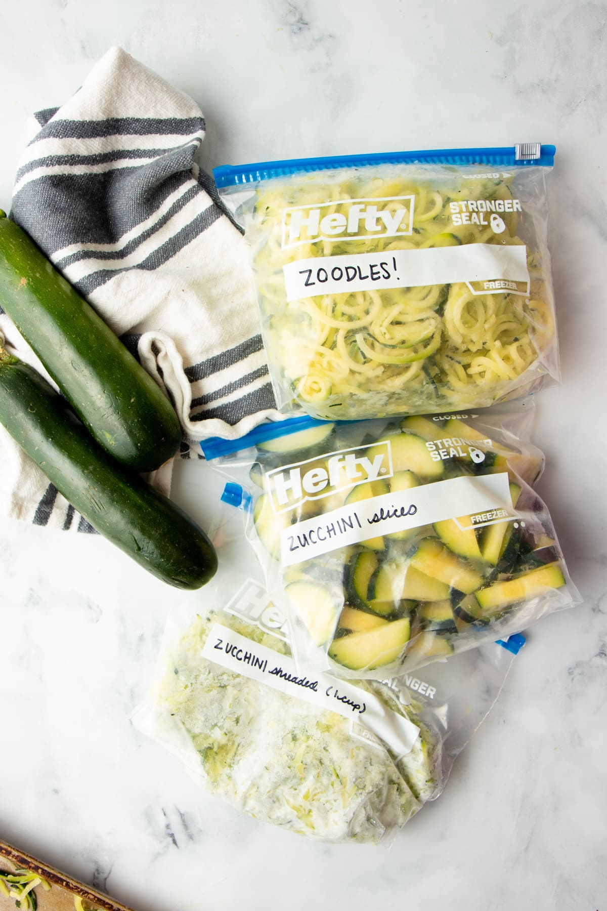 Three Hefty freezer bags filled with frozen sliced zucchini, frozen zucchini noodles, and frozen shredded zucchini.