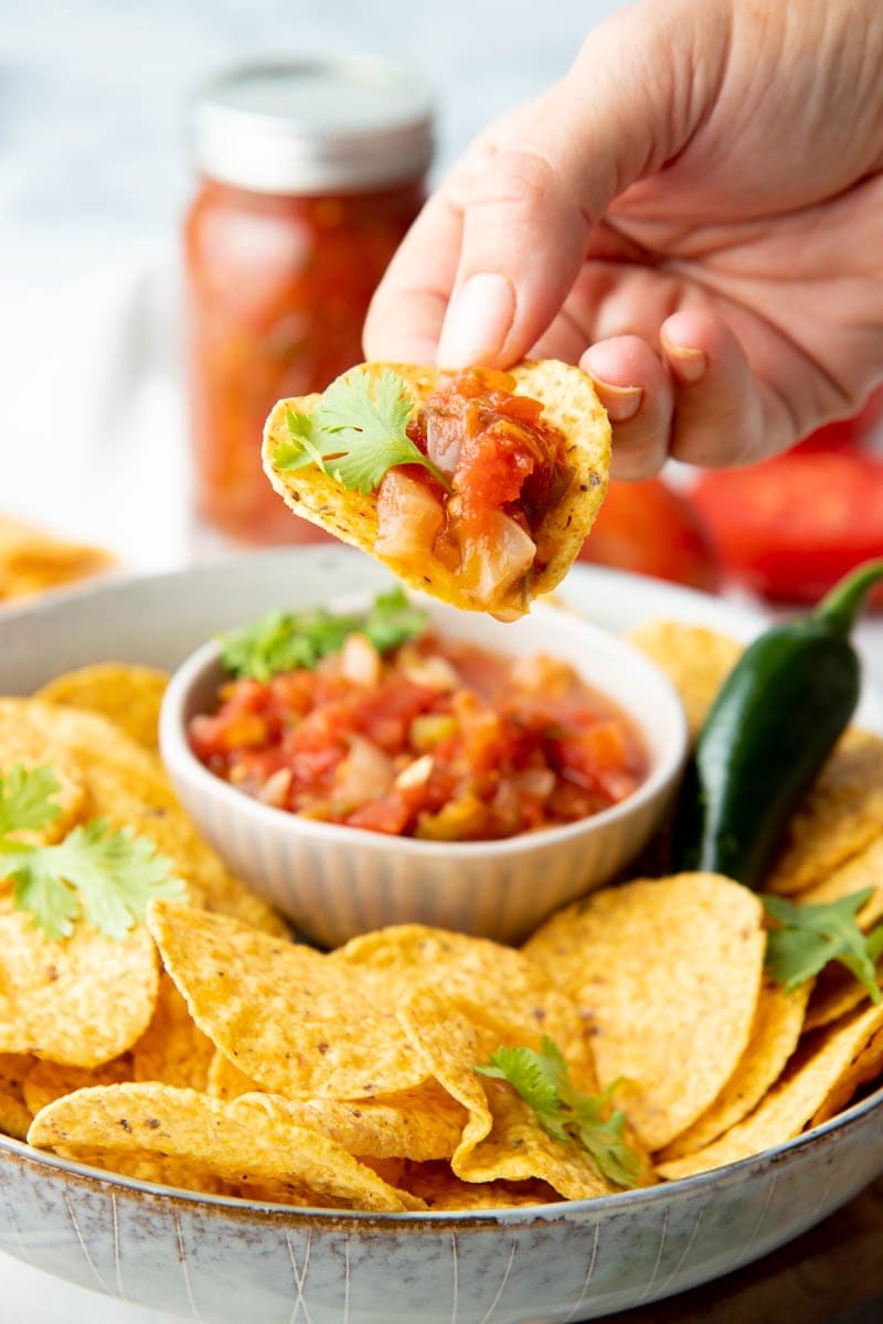 Close-up of a hand holding a tortilla chip dipped in salsa with a cilantro leaf garnish.