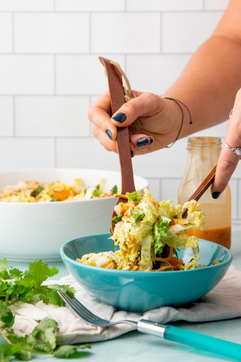 Two hands holding wooden salad servers toss a single serving of mandarin chicken salad with homemade dressing.