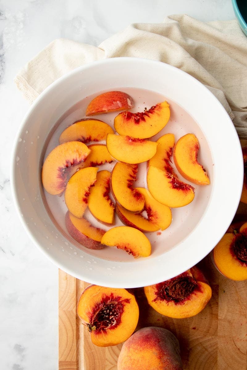 Overhead of fresh peach slices in a bowl filled with water and lemon juice to prevent browning.