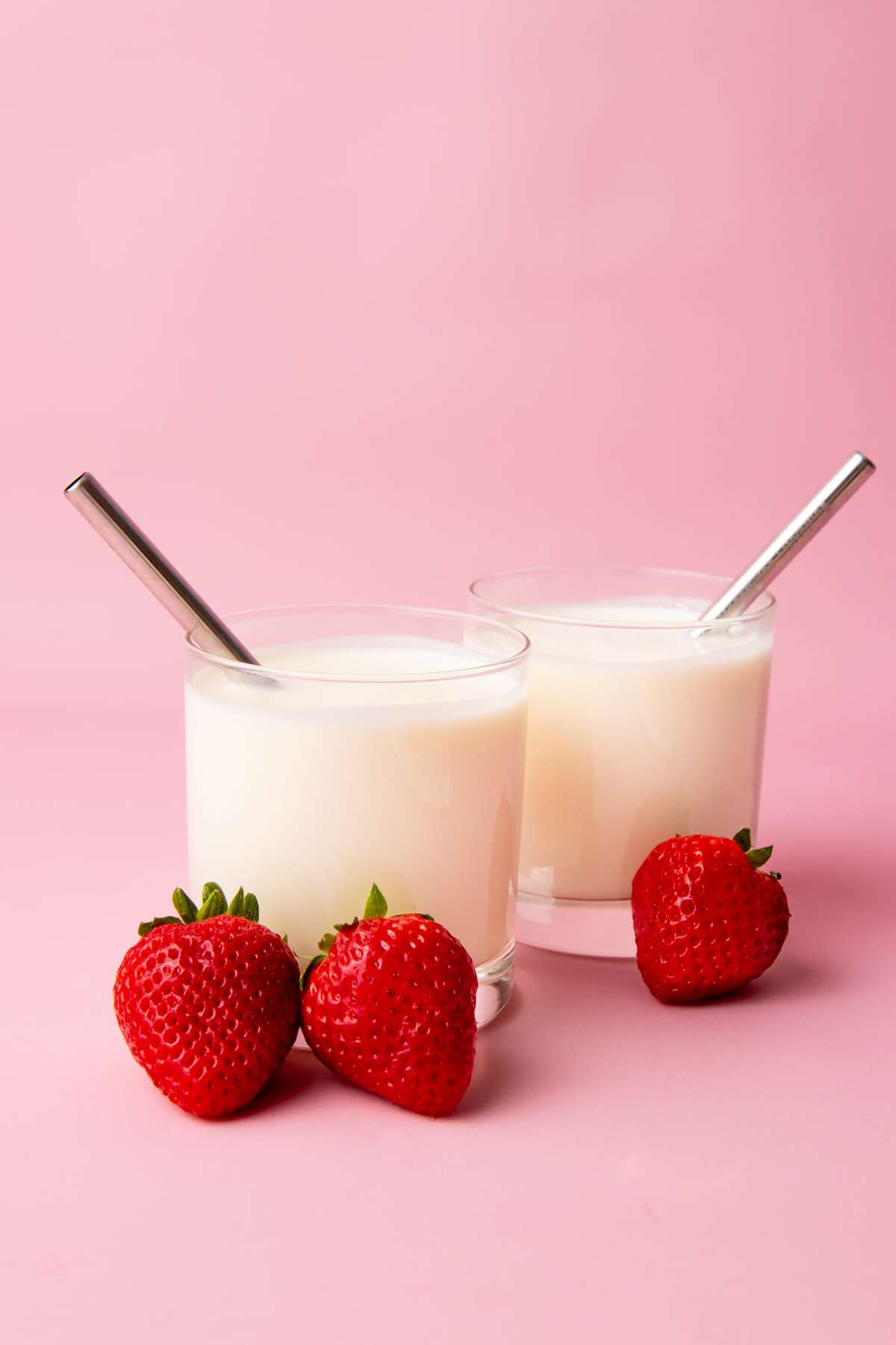 Two glass tumblers filled with homemade kefir and metal straws sit on a blush, pink background with ripe, red strawberries leaning against them.