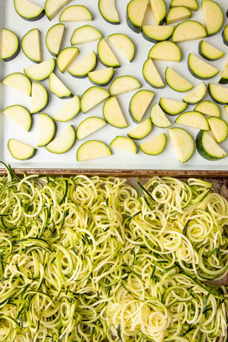 Overhead of two baking sheets, the top baking sheet filled with zucchini slices and the bottom baking sheet filled zucchini noodles.