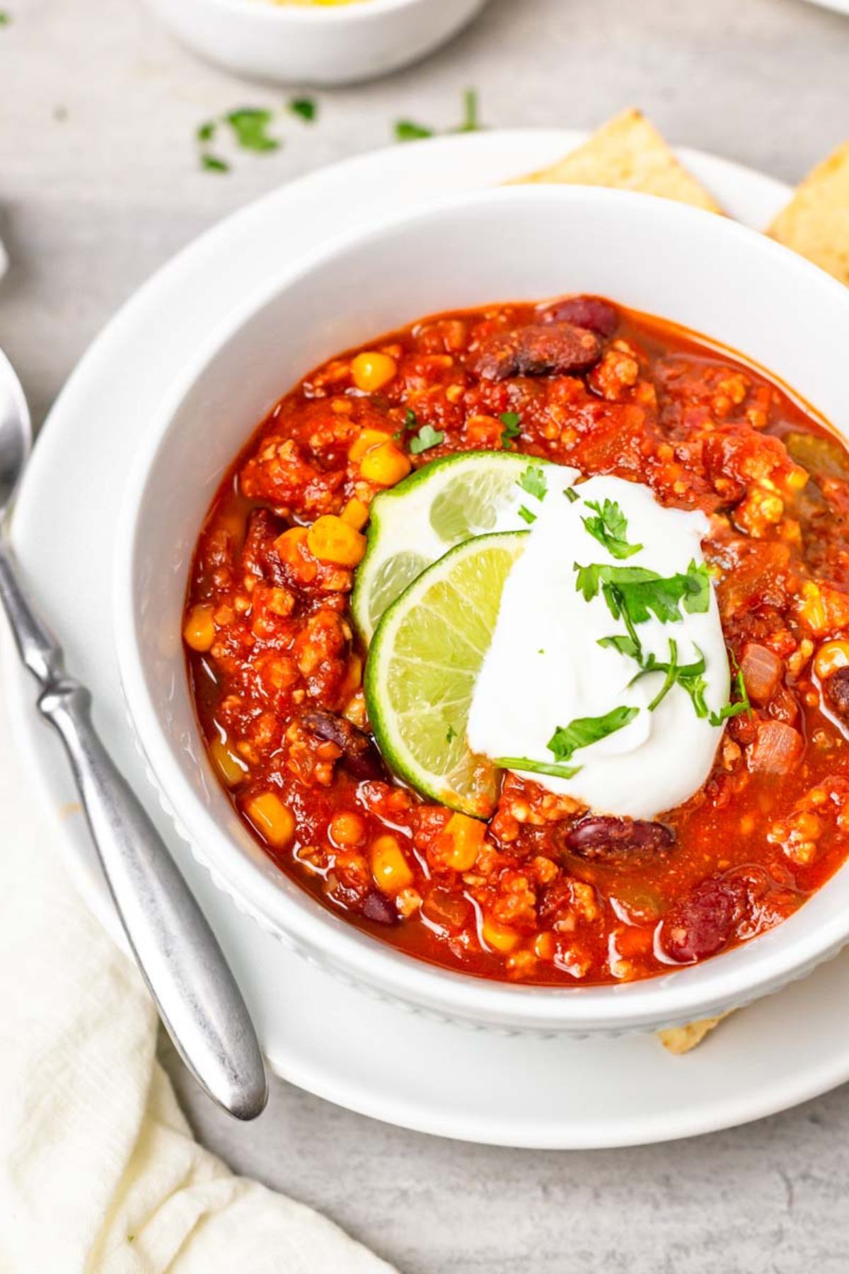 A bowl of taco chili sits on a gray countertop. The chili has lime wedges and sour cream on top.