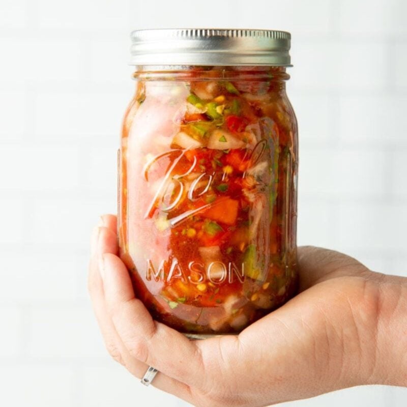 A hand holds a finished jar of canned salsa against a white tile backsplash