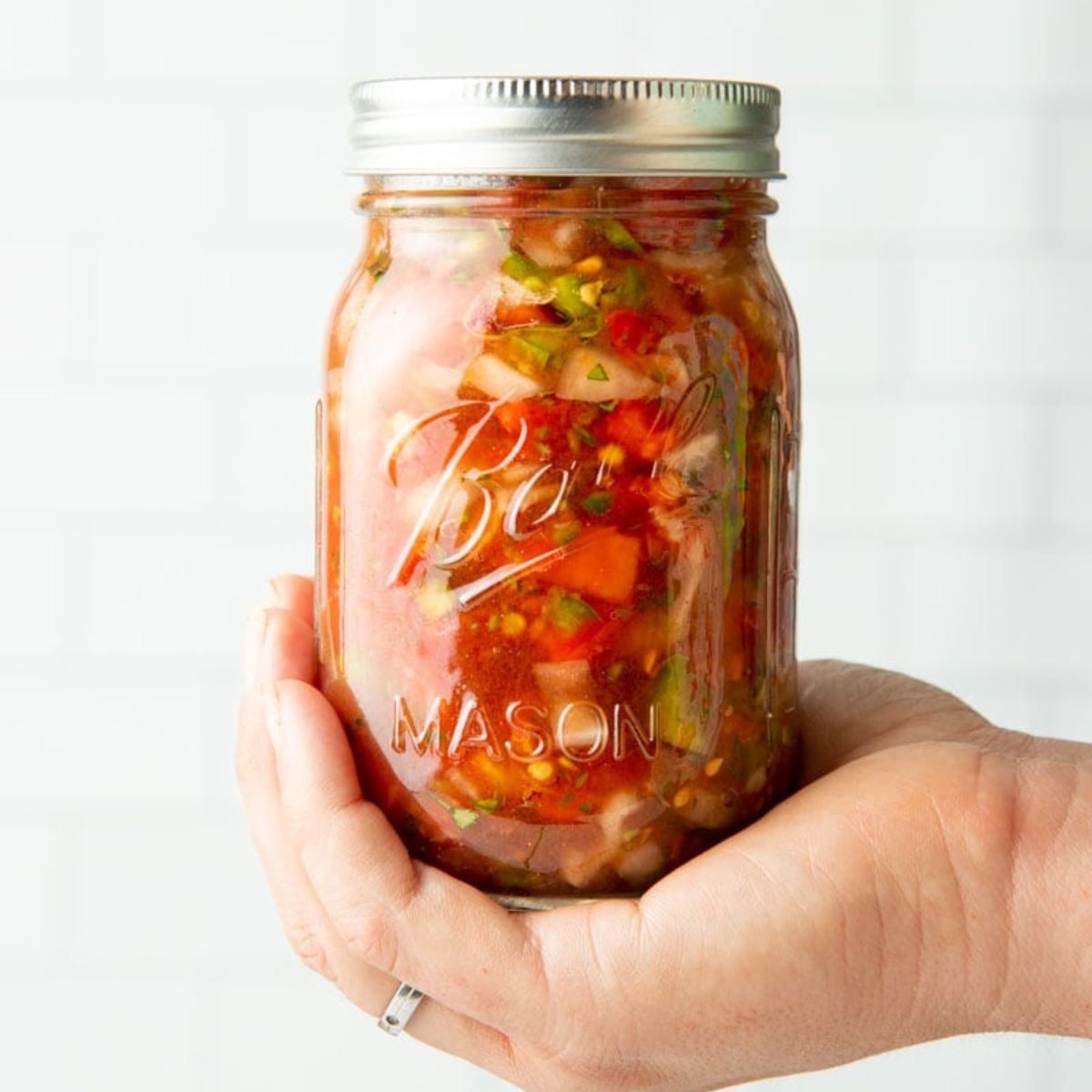 A hand holds a finished jar of canned salsa against a white tile backsplash